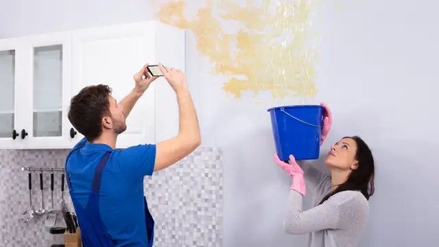 Homeowner and technician photographing a water stain on a ceiling while a bucket catches drips